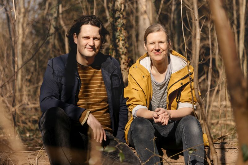 Halbfigur: Dico Kruijsse und Carolin Lange sitzen auf einem Baumstamm im Wald und  schauen in die Kamera.
